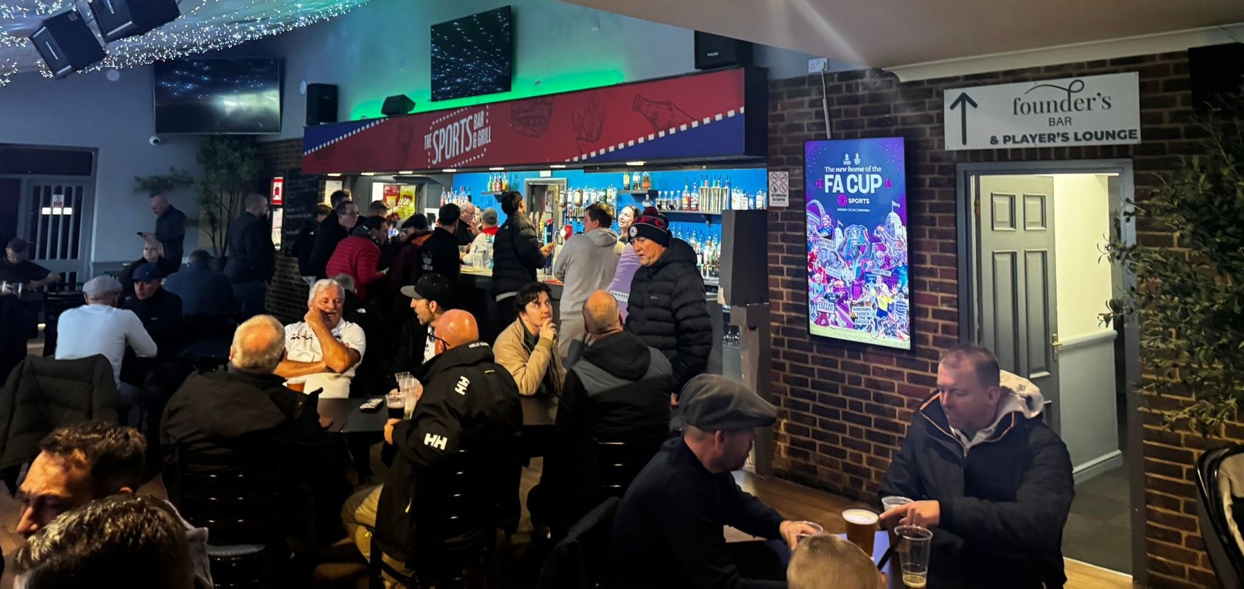 TNT Sports FA Cup creative on a portrait Grassroot Media screen beside a busy grassroots clubhouse bar
