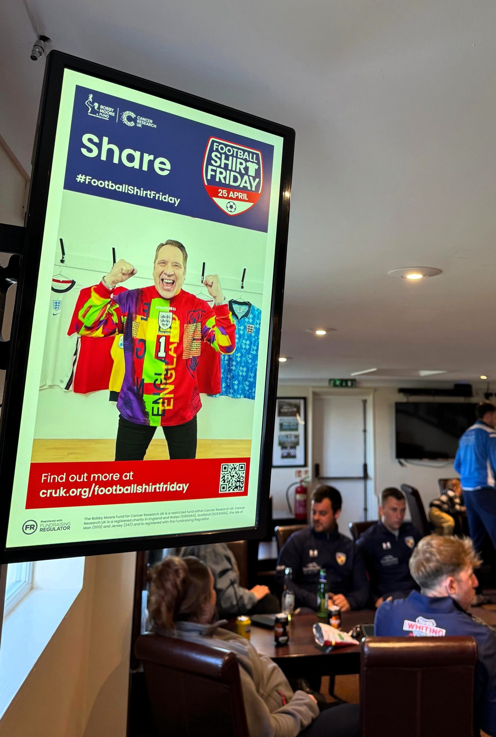 Football Shirt Friday campaign displayed on a digital screen in a grassroots rugby clubhouse, with players sitting and chatting in the background.