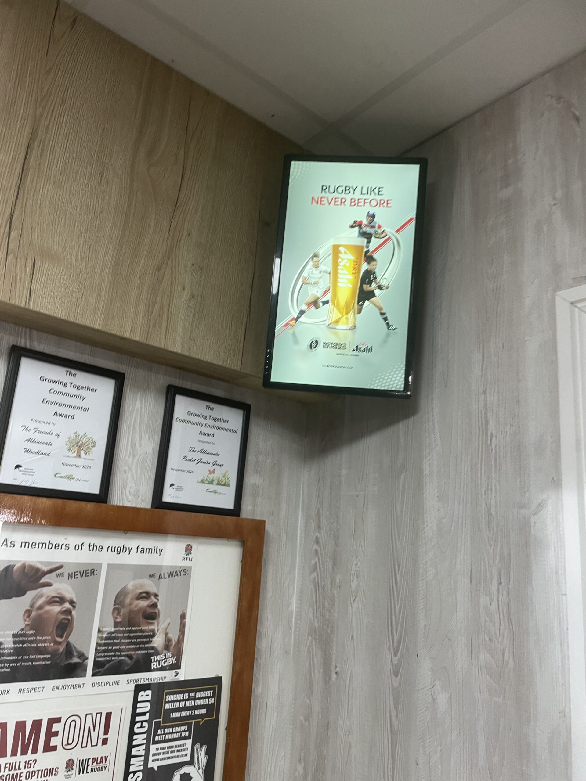 Bright rugby club interior with Asahi’s 'Rugby Like Never Before' campaign on screen, surrounded by framed award certificates and posters.