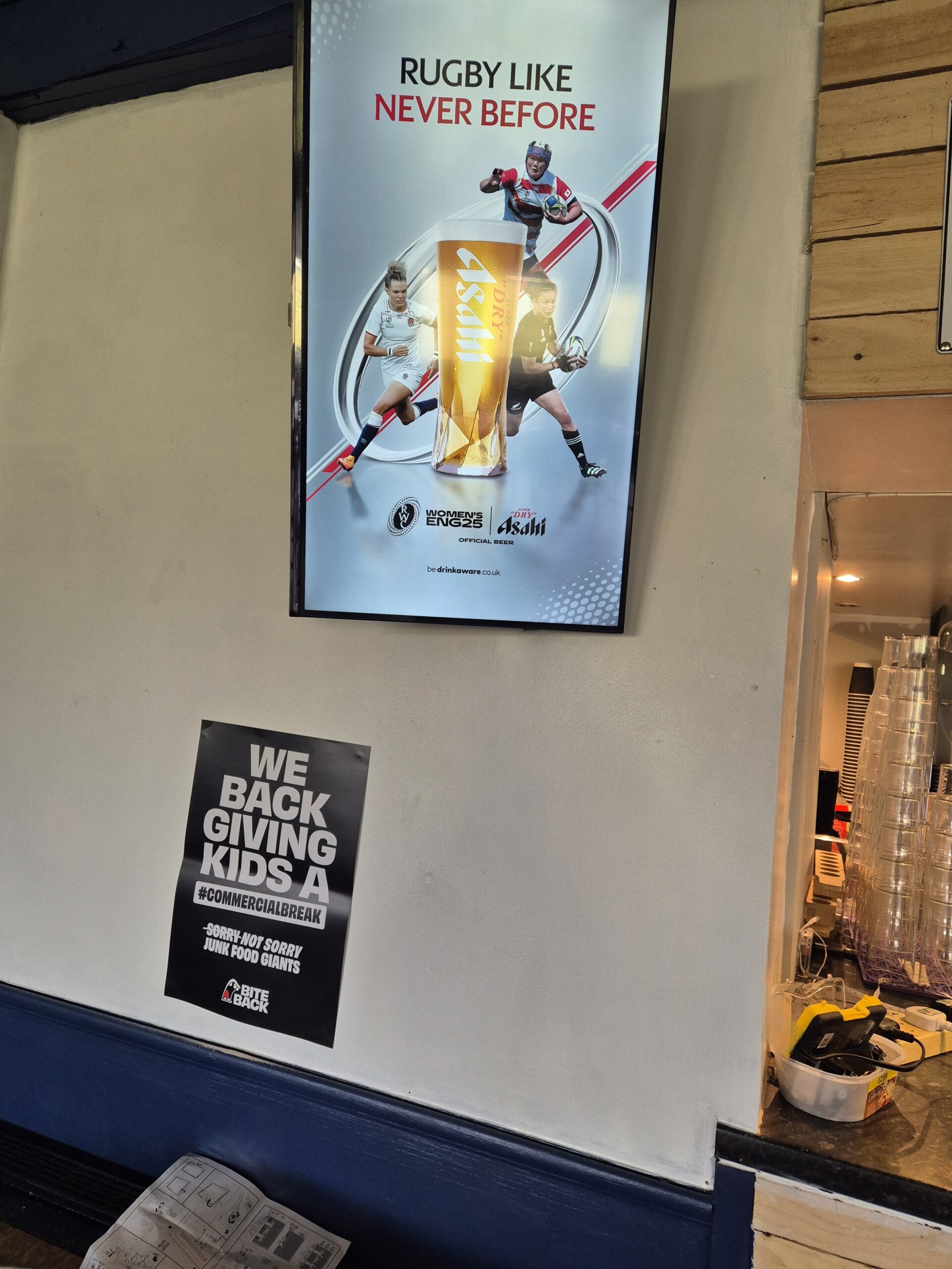 Screen above a rugby club kitchen counter showing Asahi’s Women’s Rugby World Cup ad, positioned above a Bite Back kids' health campaign poster.