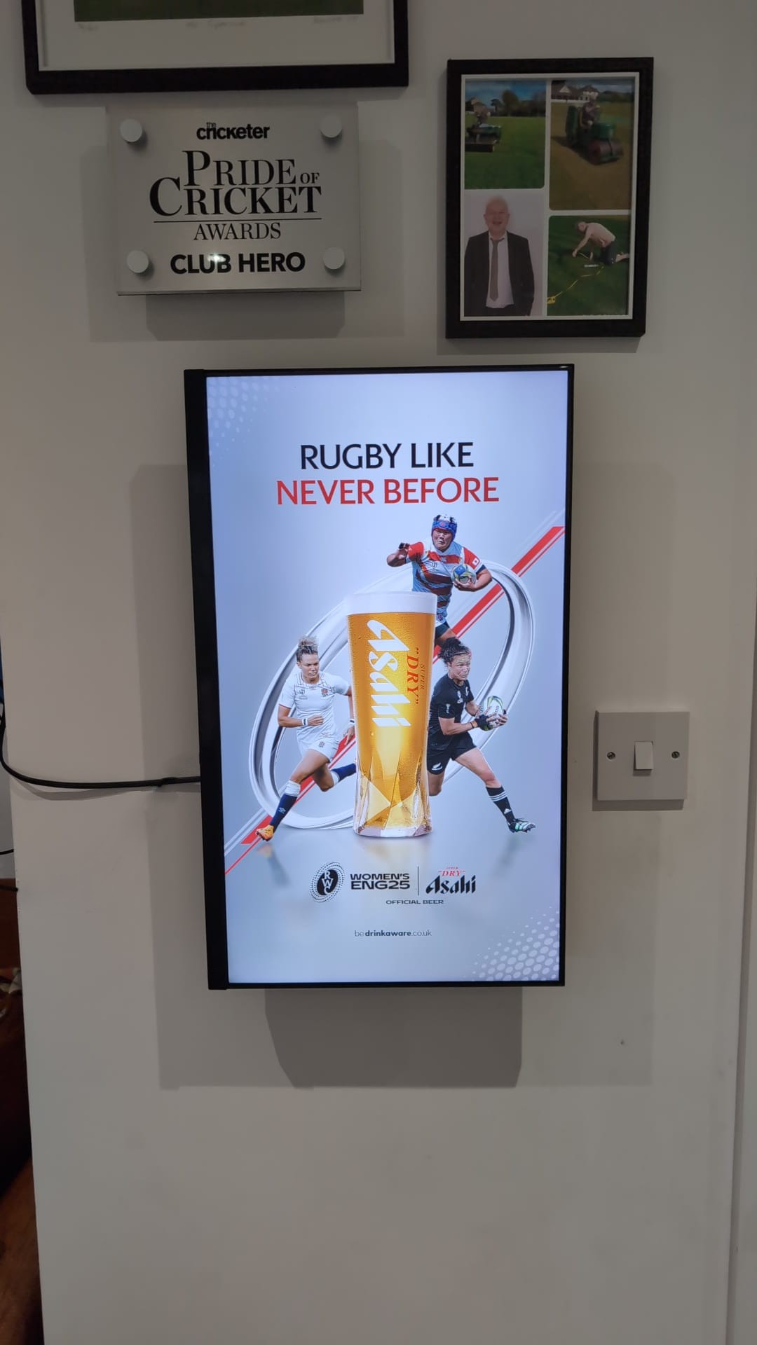 Digital screen playing Asahi's Women's Rugby World Cup advert beneath a Pride of Cricket Awards plaque and framed photographs at a sports club.
