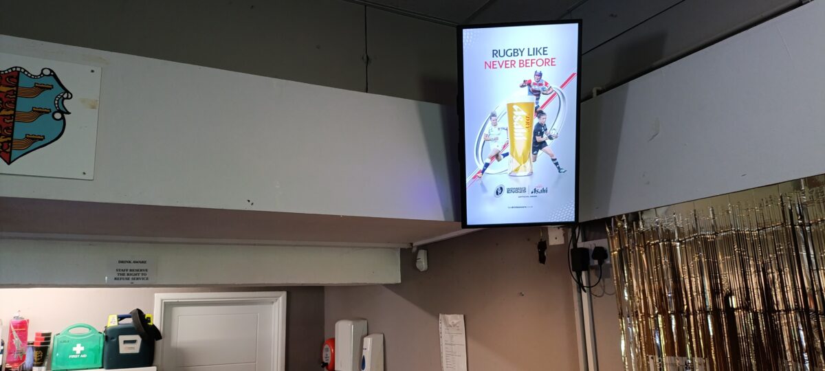 Digital screen inside a grassroots rugby clubhouse displaying Asahi’s “Rugby Like Never Before” campaign, mounted above a bar area with visible first aid kit, drinkware, and a gold foil curtain to the side.