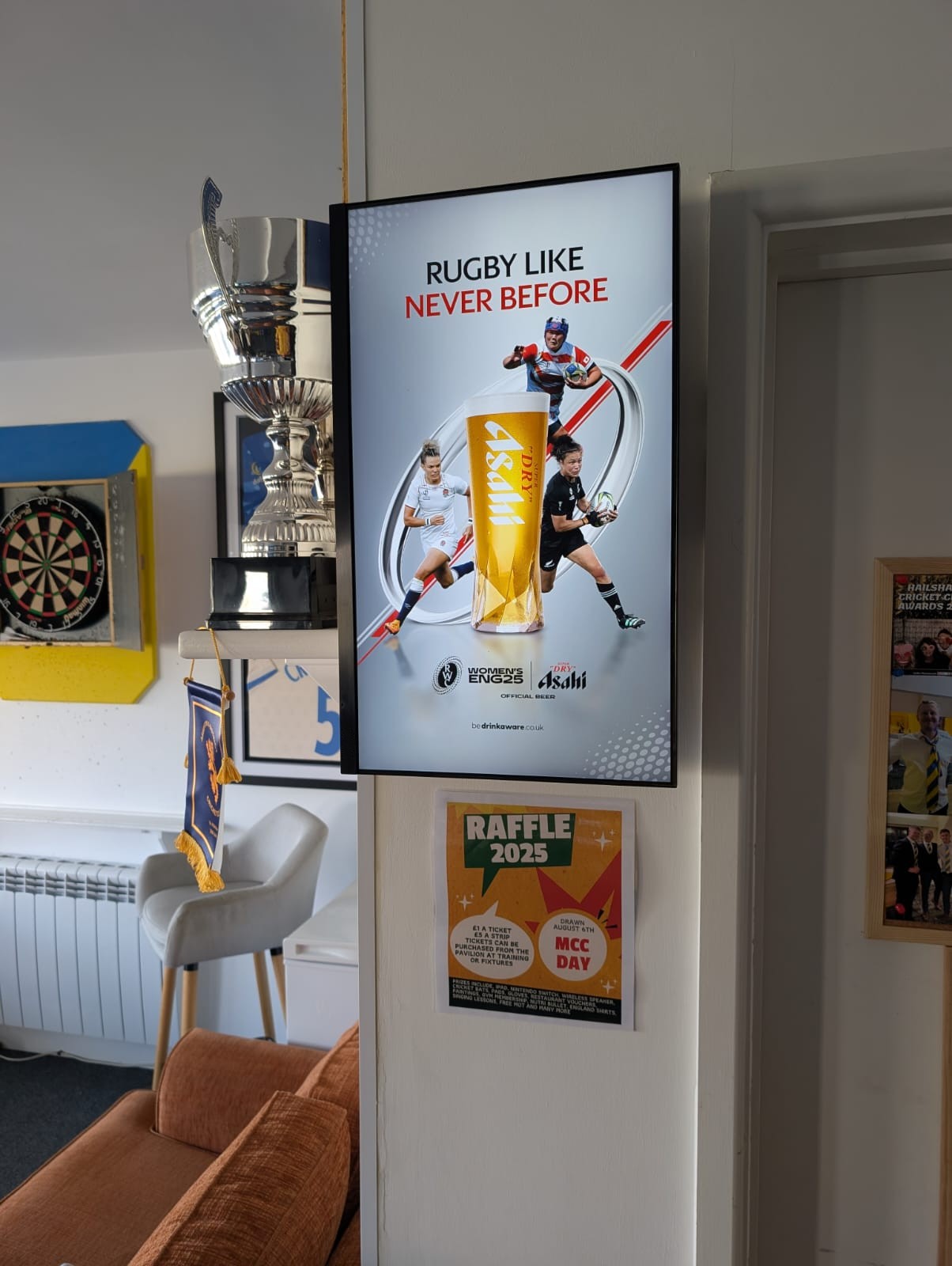 Digital screen in a grassroots sports clubhouse showing an Asahi beer rugby advert next to a dartboard, highlighting high-dwell bar advertising opportunities.