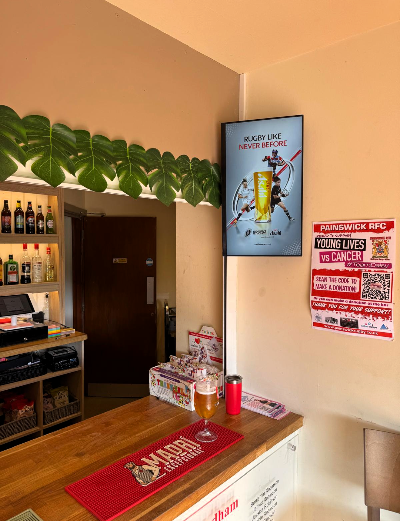 Asahi 'Rugby Like Never Before' advert shown on a digital screen behind a clubhouse bar with a Madrí beer mat and charity poster visible nearby.