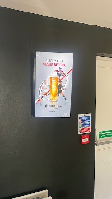 Portrait screen on a dark clubhouse wall displaying Asahi’s Women’s Rugby World Cup campaign creative, positioned above a fire alarm and safety signage.