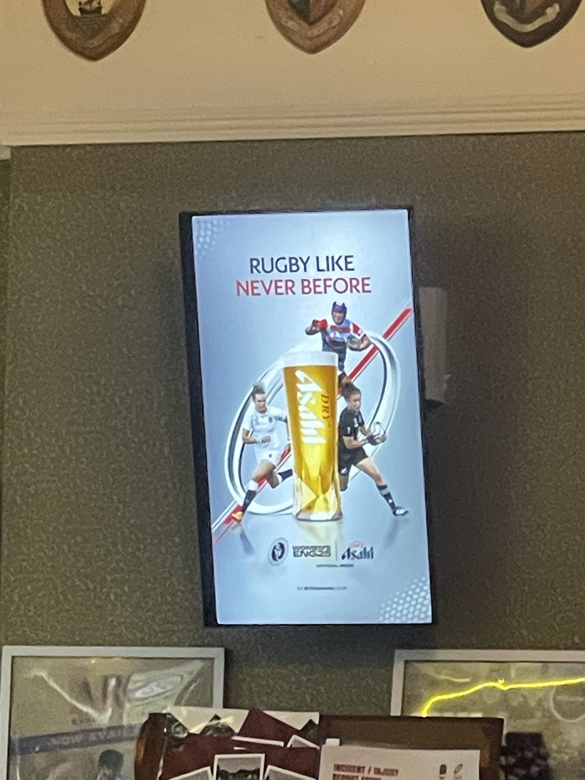 Rugby clubhouse with digital screen showing Asahi’s 'Rugby Like Never Before' ad, surrounded by team photos and wooden wall plaques.