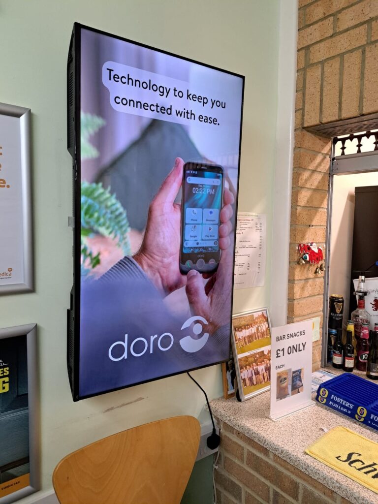 Doro mobile phone advert displayed on a portrait digital screen beside the bar in a bowls clubhouse.