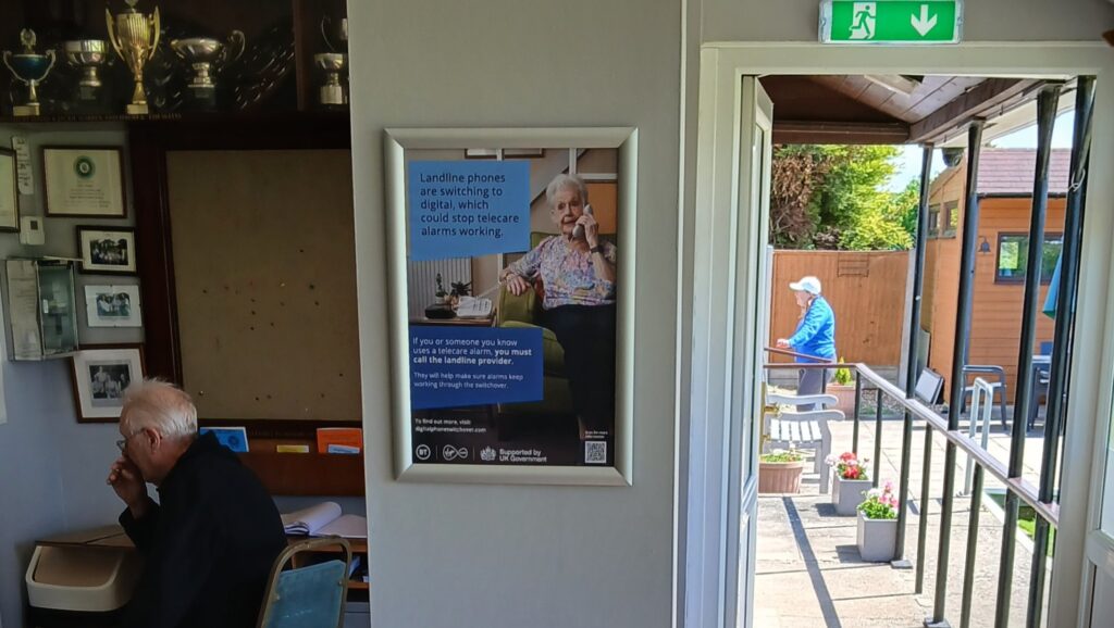 UK Government-supported telecare switchover poster displayed beside a bowls clubhouse entrance with members nearby.
