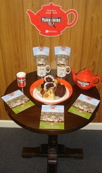 Yorkshire Tea branded teapot, mugs, cakes and campaign leaflets arranged on a display table at Reed Cricket Club.