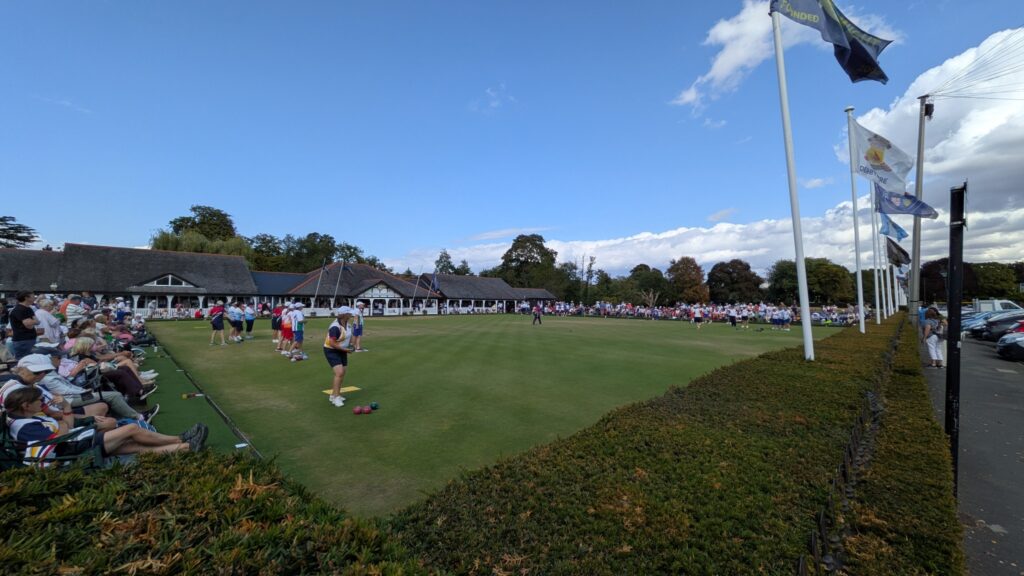 A wide view of the National Bowls Finals with spectators, players and club flags around the green