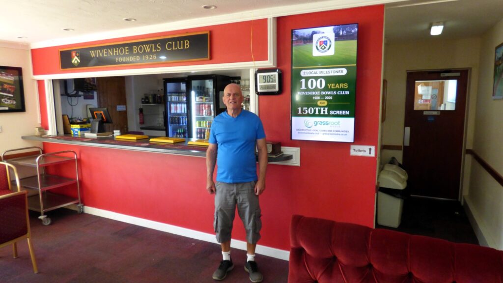 Peter Wilby standing inside Wivenhoe Bowls Club beside a digital screen marking the club’s centenary and Grassroot Media’s 150th screen